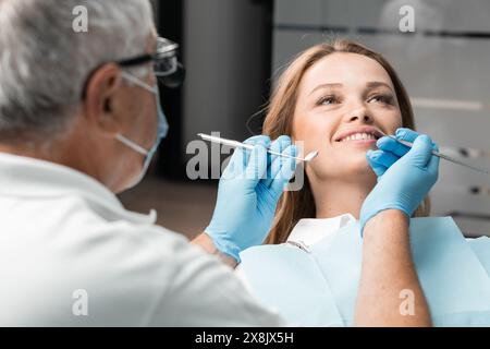 A patient is comfortably seated in the dental chair, whose bright smile adorns the office. The dentist stands nearby, ready to provide qualified Stock Photo