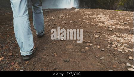 A person walks along a dirt path near a waterfall in Indonesia, Bali. The lush greenery surrounds the scene as water cascades down rocks creating a serene atmosphere. Stock Photo