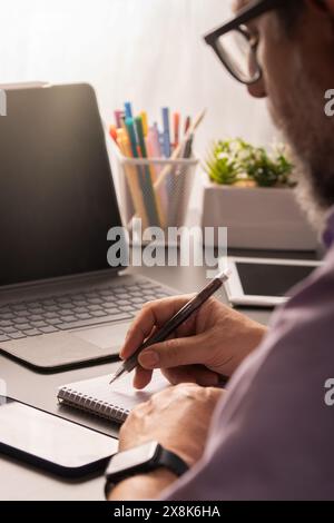 A professional man working at a desk in a home office setting, taking notes while using a laptop. The scene includes modern technology and stationery, Stock Photo