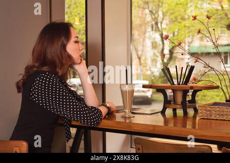 A young elegant woman in a black polka dot dress is gracefully sitting in a cozy cafe, savoring her coffee while gazing thoughtfully out the window, l Stock Photo
