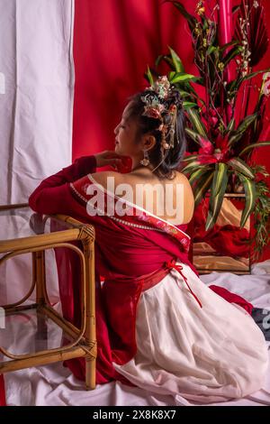 A woman in a red dress sits on a wooden chair. She has a tattoo on her back and is wearing a flower headband. The room is decorated with red and Stock Photo