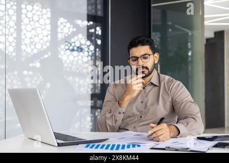 A man, looking serious and professional, focuses on work in a modern office. He analyzes data charts, takes notes, and uses a laptop at his desk, showing concentration in his tasks Stock Photo
