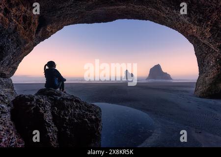 Female tourist sitting on rock in stone cave with Archway islands on Wharariki beach in the morning at Cape Farewell, New Zealand Stock Photo