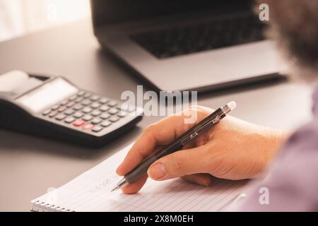 A person writing notes on a notepad with a pen, accompanied by a calculator and laptop on a home office desk. The setup highlights financial planning Stock Photo