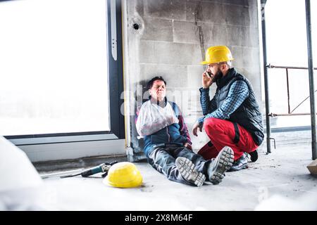 Colleague calling ambulance and giving first aid to injured worker, in pain after accident at construction site Concept of occupational safety and Stock Photo