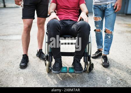 Close up of legs of young man sitting in wheelchair, friends standing by him. Male friendship. Stock Photo