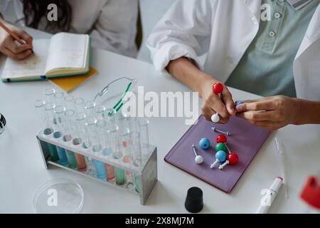 High angle view of unrecognizable kids sitting at desk in classroom assembling chemical structure model and making notes during lesson Stock Photo