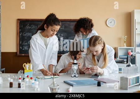 Multi-ethnic group of boys and girls wearing lab coats using microscope while doing experiment in modern Chemistry class Stock Photo