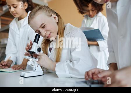 Selective focus shot of Caucasian schoolgirl sitting at desk in classroom using microscope during Chemistry lesson Stock Photo