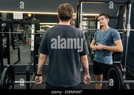 Rear view of beginner sportsman exercising at gym support under supervision of coach. Athlete hard time doing barbell press during weightlifting Stock Photo