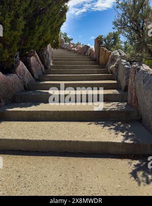 Cement steps lined with rocks & shadows lead uphill showing distant bright blue sky with white clouds. Rocks in Cody, Wyoming, used as railing wall. Stock Photo