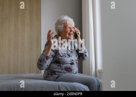 Senior woman at home, phone calling, sitting on bed. Older woman living alone, enjoying peaceful weekend day. Stock Photo