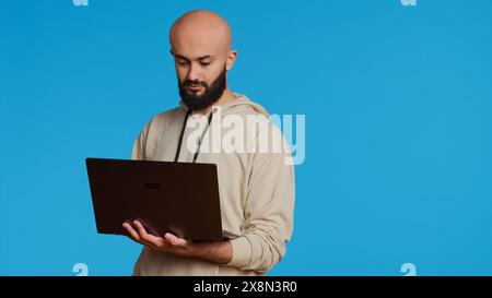 Middle eastern man works with cyber security on laptop, trying to do some software programming and report bugs. Young adult operating as a system developer in studio. Camera 2. Handheld shot. Stock Photo