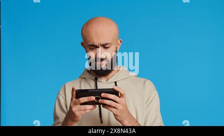 Middle eastern person playing mobile video games on app, standing over blue background and enjoying online tournament using mobile phone. Man having fun with gaming contest. Camera 2. Stock Photo