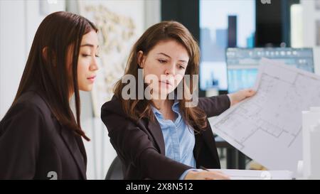 Designers analyze 3D building models to generate specifications, examining maquettes to develop production blueprints. Team of architects focus on industrial drafts in shared workspace location. Stock Photo