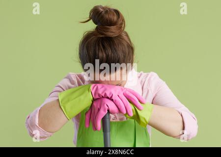 Spring cleaning. sad modern middle aged woman in green apron and rubber gloves isolated on green with mop and headache. Stock Photo