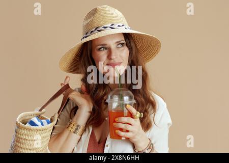 Beach vacation. happy stylish woman in white blouse and shorts against beige background with straw bag, carrot juice and straw hat. Stock Photo