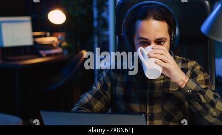 Happy man drinking coffee and writing emails at home office desk while listening music. Smiling remote worker doing b2b networking over internet, wearing headphones, enjoying hot beverage, camera A Stock Photo
