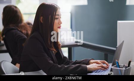 Asian architect translating floor plan measurements on laptop, studying architectural blueprints and helping contractors with dimensions. Real estate agency worker overseeing project. Handheld shot. Stock Photo