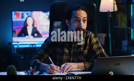 Remote employee working at home office desk, reading information on laptop and writing in notebook. Freelancer noting down ideas on paper using pen while teleworking, camera A Stock Photo