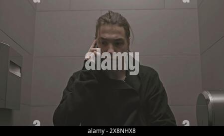 Young man sits on toilet and talks on phone. Stock. Man in black walks into toilet stall and talks on phone. Young man is talking aggressively on Stock Photo