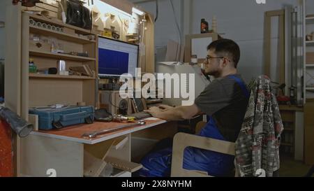 Man is sitting on computer in carpentry workshop. Creative. Man with computer drawings for processing wooden products. Workshop for woodworking and Stock Photo
