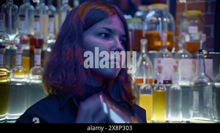 Young female bartender at nightclub. Media. Female bartender shaker drink at bar. Bartender prepares drink on background of many bottles of alcohol in Stock Photo