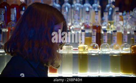 Young female bartender at nightclub. Media. Female bartender shaker drink at bar. Bartender prepares drink on background of many bottles of alcohol in Stock Photo