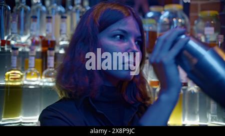 Young female bartender at nightclub. Media. Female bartender shaker drink at bar. Bartender prepares drink on background of many bottles of alcohol in Stock Photo