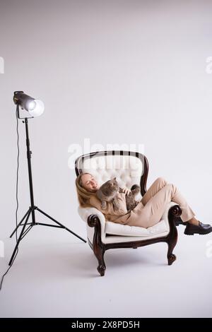Female blogger, blonde on chair with domestic cat scottish straight in white photo studio. Dressed in a formal beige trouser suit. Vertical Stock Photo