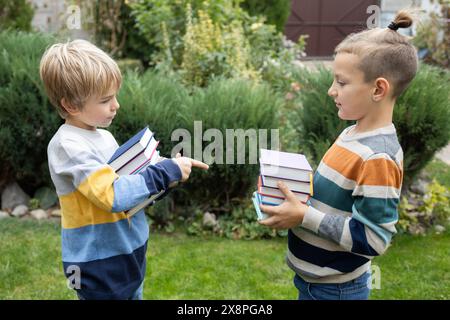 Two brothers or boy friends stand opposite each other hold stacks of books in their hands. lovely schoolchildren aged 7-8 years old, interested in lea Stock Photo