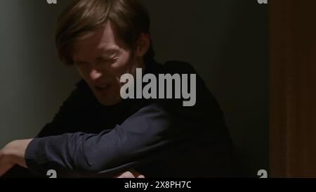 Frustrated young man squatting down against the wall and wrinkling his face. Stock clip. Struggling with unbearable headache. Stock Photo