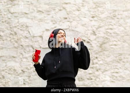 Portrait of a young woman wearing a black clothes and headphones is joyfully singing along to music, with a bright and happy expression on her face. Stock Photo