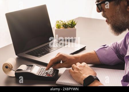 A businessman working in a home office, using a calculator and laptop for budget calculations. The setup emphasizes financial planning and an organize Stock Photo