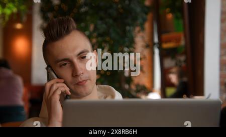 Young man is working on laptop in cafe and talking on phone. Stock footage. Young freelancer is working on laptop and answering calls. Remote work Stock Photo