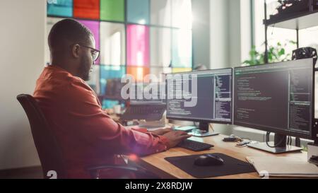 African American Man Coding on Desktop Computer with Professional Multiple Monitors Setup in Creative Office. Male Software Engineer Developing Mobile Application for Innovative Start-Up. Stock Photo