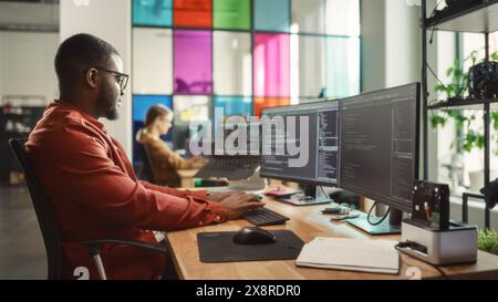 African American Man Coding on Desktop Computer with Professional Multiple Monitors Setup in Creative Office. Male Software Engineer Developing Mobile Application for Innovative Start-Up Company. Stock Photo