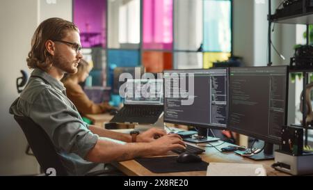 Young Caucasian Man Coding on Personal Computer and Laptop Set Up In Stylish Office. Professional Programmer Developing Innovative AI Software in Technological Start-Up, Using Multiple Displays. Stock Photo