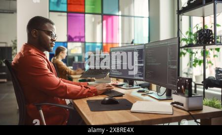Black Man Writing Lines of Code On Desktop PC With Two Monitors and a Laptop Aside in Stylish Office. Professional Male Developer Programming Artificial Intelligence Software for Start-Up Company. Stock Photo