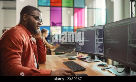 Black Man Writing Lines of Code On Desktop PC With Two Monitors and a Laptop in Stylish Office. Professional Male Developer Programming Artificial Intelligence Software for Start-Up Company. Stock Photo