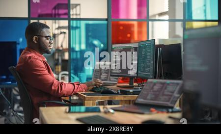 African American Man Writing Lines Of Code On Desktop Computer With Multiple Monitors and Laptop in Creative Office. Male Data Scientist Working on Innovative Online Service For Start-up Company. Stock Photo