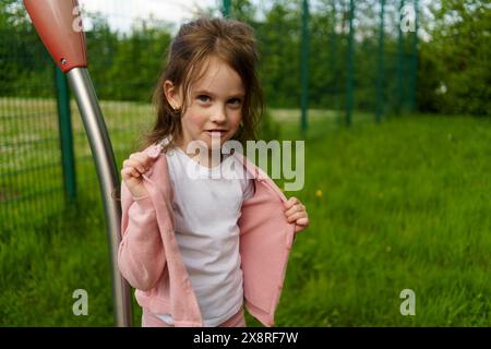 A little smiling, beautiful girl takes off a sweater on a summer playground. Stock Photo