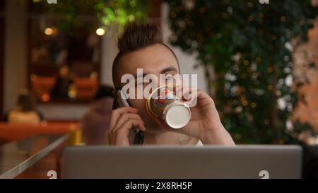 Young man is working on laptop in cafe and talking on phone. Stock footage. Young freelancer is working on laptop and answering calls. Remote work Stock Photo