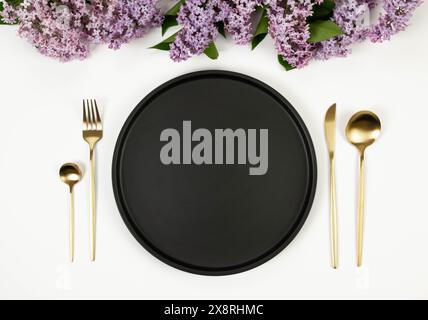 Top view of purple lilac flowers on white background. Table setting, black plate, gold cutlery. Spring wallpaper, flat lay, copy space. Stock Photo