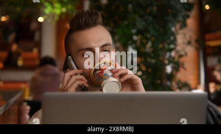 Young man is working on laptop in cafe and talking on phone. Stock footage. Young freelancer is working on laptop and answering calls. Remote work Stock Photo