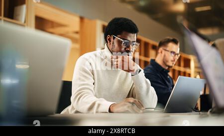 Portrait of Black Creative Young Man Arriving at Office for Work Office and Working on Laptop. Supervisor Smiling While Answering Email Questions From Colleagues. People Working in the Background Stock Photo