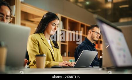 Group of Diverse Creative Team Members Using Laptops in an active Contemporary Office. Young Female Media Relations Manager Working on Brand Innovation Strategy and Collaborating with Colleagues. Stock Photo