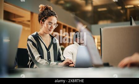 Diverse Group of Creative Colleagues Working on Laptops in Modern Office. Hispanic Female Graphics Designer Smiling and Typing. Colleagues Working on Projects in Background. Stock Photo
