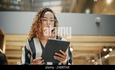 Portrait of a Powerful Female Leader Checking Meeting Briefing on her Tablet and Walking in Busy Corporate Office. Confident Hispanic Team Leader Preparing for a Presentation. Low Angle Shot Stock Photo