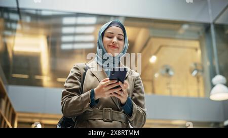 Confident Female Team Manager wearing Hijab Answering Emails on Smartphone After Receiving Good Performance Report. Portrait of a Young Muslim Woman Walking in a Hallway of an Office and Smiling. Stock Photo